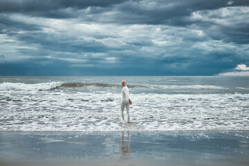 Hairless girl with alopecia in white futuristic suit goes to cold restless sea on sandy beach,...