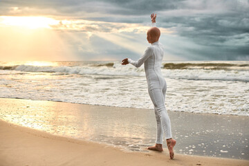 Young hairless ballerina with alopecia in white futuristic suit dancing on seashore at sunset sea,...