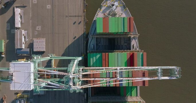Birds eye top down view of cranes unloading a docked container ship with trucks driving alongside