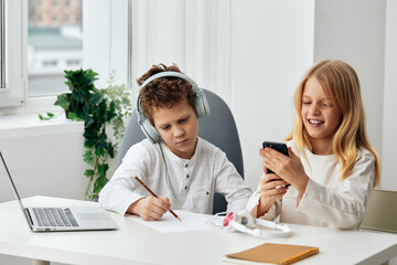 Happy Caucasian boy and girl studying at home with their laptops, headphones, and phones for elearning They are sitting at a table in the living room, engrossed in their online lessons The room is