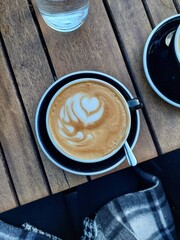 Top view of a cup of cappuccino on a wooden table in a black cup with heart-shaped coffee art with a hint of the woman's legs under the table and her black and white coat