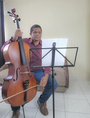 Music student playing cello and guitar at home, sitting, with music stand and sheet music.