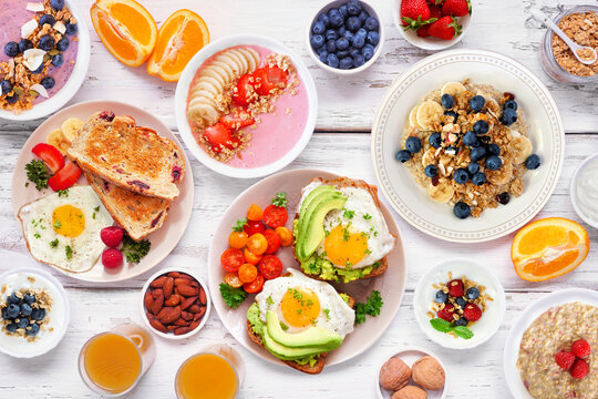 Healthy breakfast or brunch table scene on a white wood background. Overhead view. Avocado toast, smoothie bowls, oats, yogurt and assorted nutritious foods.