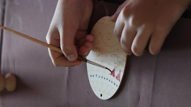 Vertical video. Close-up of little girl painting wooden Easter egg in the kitchen
