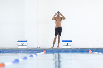 Young biracial male athlete swimmer adjusts his goggles at the poolside