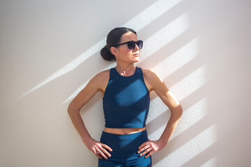 sporty woman standing against a wall in the sun, wearing sunglasses.