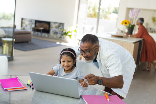 Biracial grandfather helps a young granddaughter with schoolwork at home using a laptop