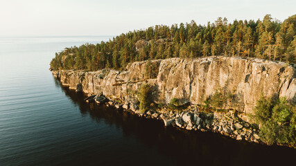 A majestic cliffside adorned with tall pines looms over the calm sea, captured in the warm light of a setting sun. The rugged beauty of the landscape speaks to the enduring nature of the coast. 