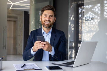 Portrait of a young businessman sitting in a suit at a desk inside the office, holding a mobile phone, smiling and looking at the camera
