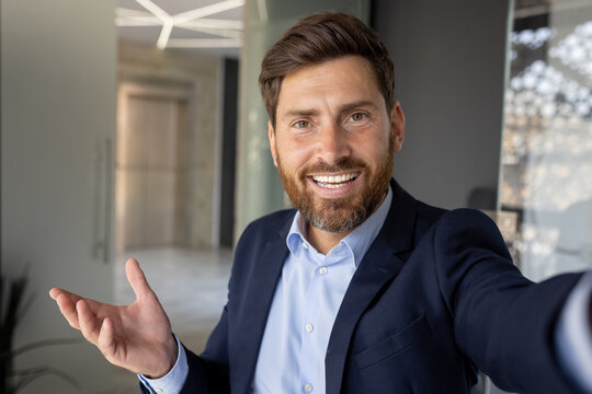 Remote video call conversation in the office. A young businessman is standing in a suit and holding a phone, looking at the camera and smiling. Close-up photo
