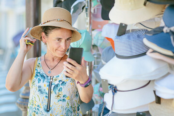 Mature woman choosing and trying on straw hat in street shop in resort town. Summer fashion and...