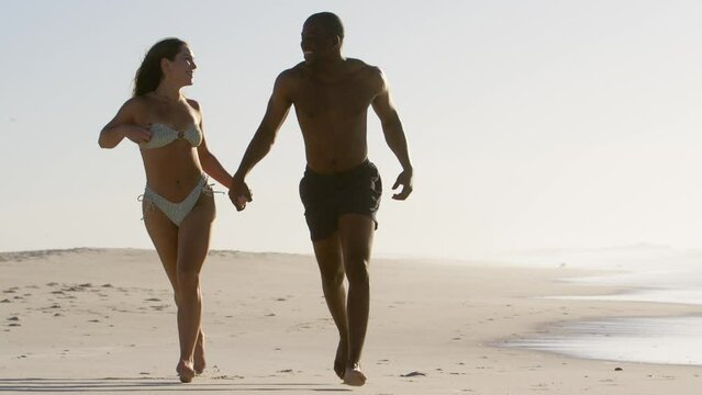 Full length shot of loving young couple in swimwear holding hands and running along beach in South Africa - shot in slow motion