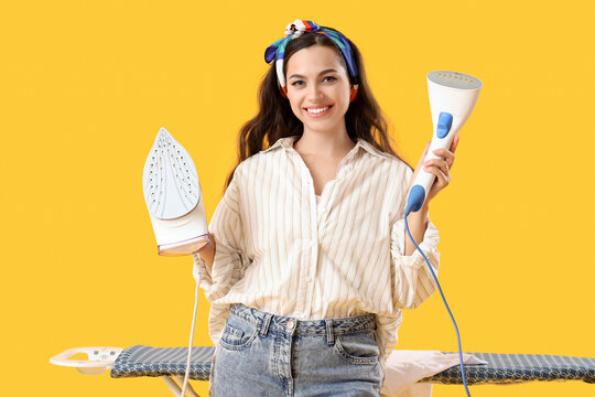 Pretty young woman with iron and garment steamer standing near ironing board on yellow background