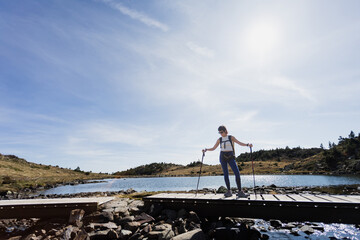 Person in middle of Pyrenees mountain hiking with walking sticks crossing bridge and lake in background