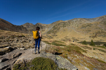 Person in middle of Pyrenees mountain hiking with walking sticks crossing bridge and lake in background