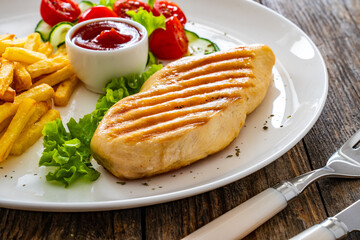 Grilled chicken breast with fries and fresh vegetables on wooden table
