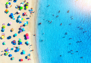 Aerial view of colorful umbrellas on white sandy beach, swimming people in blue sea on summer sunny...