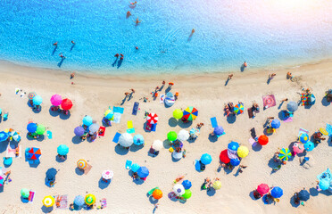 Aerial view of colorful umbrellas on white sandy beach, swimming people in blue sea at sunset in summer. Resort in Sardinia, Italy. Tropical seaside with turquoise water. Travel and vacation. Top view