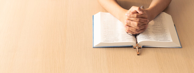 Cropped image of praying male hand holding cross on holy bible book at wooden table. Top view....