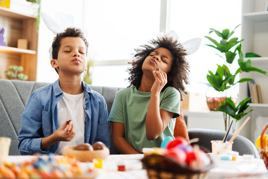 Smiling African American kids wearing bunny ears eating tasty chocolate eggs celebration Easter at home