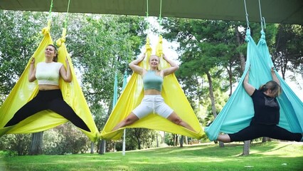 Woman yoga aero air asana in hammock in public park. Training fresh air. Stretching, meditating and mental wellbeing for females adults of middle age. Health body maintenance. Open air activities - Powered by Adobe