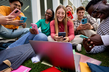 Group of cheerful young multiracial university students immersed on their mobile cell, technology addicted friends sitting outside university campus. Smiling gen z people concentrated using smartphone