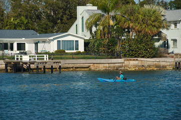 Fototapeta na wymiar Man in blue kyack lower right of water with seawall, houses and green trees in the background. On a sunny day Tampa Bay Florida later afternoon. Horizontal shot. Medium wide shot.