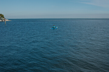 Naklejka premium Man in blue kayak in the middle with wood post and seawall on the left looking out to Tampa Bay and blue calm water on a sunny afternoon. Horizontal shot with room for copy. blue sky in the back. 