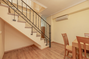 Interior staircase of a duplex residential house with wrought metal railings, cherry-colored wooden furniture, empty space under the stairs and wooden flooring