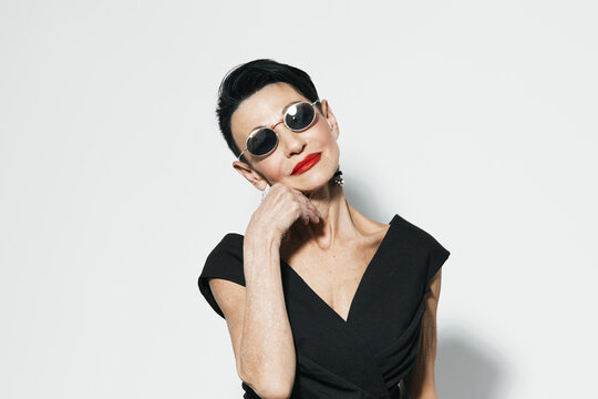 Fashionable woman in black dress and sunglasses posing in front of white wall for camera shoot