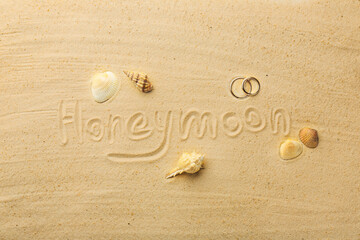 Word Honeymoon written on sand, two golden rings and seashells, flat lay
