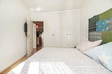 Bedroom with custom built-in wardrobe with white lacquered wooden doors, varnished oak floor and bed with small flower printed bedspread