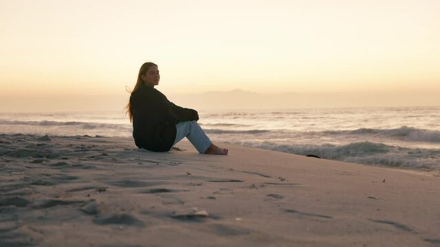 Camera tracks across shot of casually dressed young woman sitting on sand watching beautiful sunrise morning over beach and sea in South Africa - shot in slow motion
