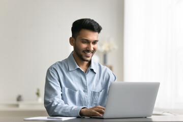 Smiling young Indian man working or studying using wireless computer, typing text on laptop keyboard, share messages, solve business by e-mailing to client, read good news. Modern tech usage, workflow
