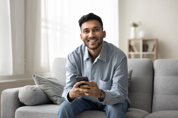 Smiling young Indian man spend free time on internet sitting on couch with smartphone. Portrait of...