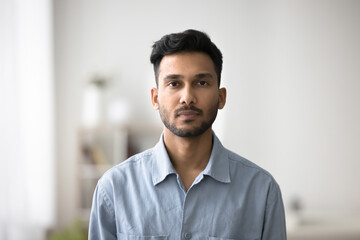 Serious Indian guy in casual blue shirt posing for camera standing alone in modern living room....