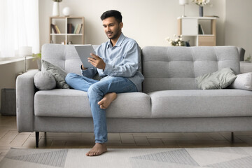 Indian man rest on couch in living room look at digital tablet screen, read e-mail, chatting,...