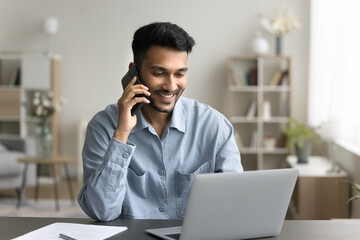 Smiling Indian guy provide information to client by phonecall, sit at desk look at laptop screen,...