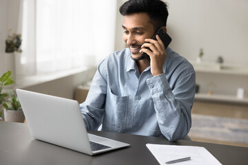 Smiling young Indian man freelancer talking on smartphone seated at desk with laptop, working at home, having business call, make phonecall to customer services or client, enjoy pleasant conversation