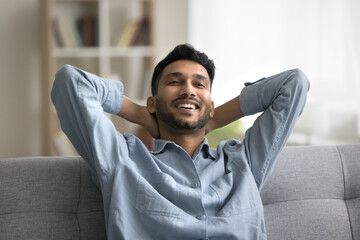 Satisfied Indian guy enjoy pleasant relaxation alone indoor, leaned on comfortable sofa, put hands...