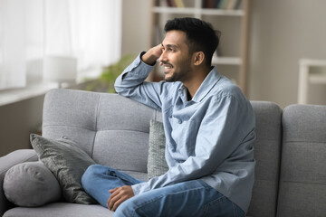 Satisfied handsome Indian guy relaxing seated on cozy sofa smiling looking out window admire view, enjoy pleasant daydreams on carefree weekend at own or rented apartment. Accommodation, day-off, rest