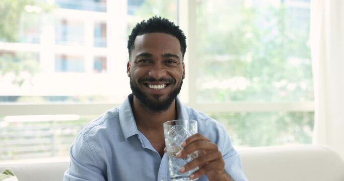 Positive thirsty handsome young African man drinking fresh clear water, holding glass, turning look at camera, posing with toothy smile, holding glass, promoting health care, hydration balance