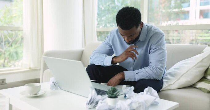 Tired overworked African freelance worker sitting at laptop, leaning head on hand, resisting sleeping, struggling to work hard, looking at computer screen at table with crumpled papers
