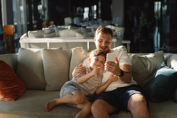 Father and son sit relax on couch in hotel hall have fun using cellphone together. Boy and dad makes selfie phone. Father and little boy child rest on sofa and watching funny video on smartphone.