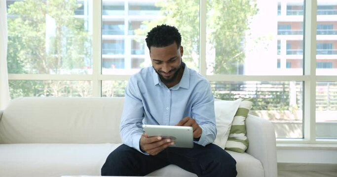 Happy attractive young African entrepreneur man using Internet technology, smart home application on digital tablet, sitting on cozy couch, typing on touchscreen, enjoying leisure, smiling