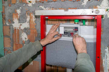 A mature  experienced plumber installing the frame of a wall-mounted toilet on  brick wall in a construction site. Builders apartment improvement or restoration works.