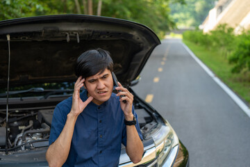 Young asian man has broken down car on the road she feeling serious and stressed. Look for someone help. Using smartphone Call Emergency assistance or insurance service.