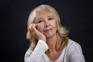 Portrait of a beautiful elderly woman. A smiling blonde sits with her hand resting her cheek. Happy maturity. Close-up. Black background.