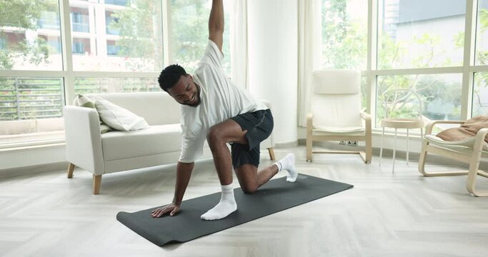 Positive athletic young African man exercising at home, twisting core on mat, stretching hips in lunge, doing yoga alone, training body, enjoying sport, fitness activity, active lifestyle