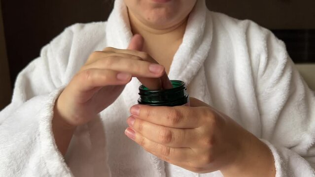 4K video. Young white woman in bathrobe takes out moisturizing cosmetic cream from green glass jar closeup in hotel bedroom. Skin protection during cold season. Cosmetic procedure in spa salon